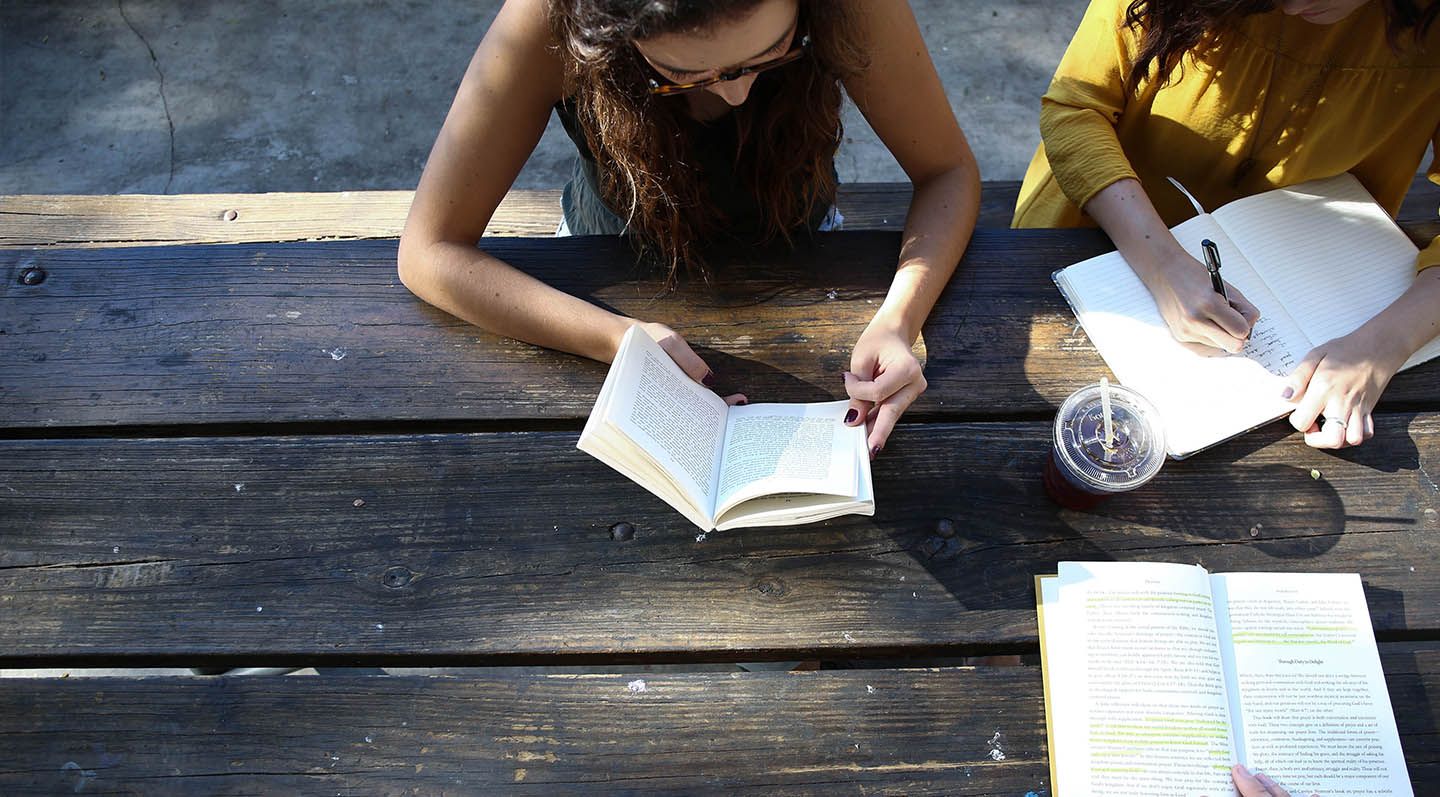 two women seated at a wooden tabel. One is reading from a book, the other is writing in a notebook